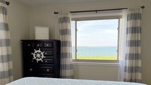 Un dormitorio con cama, cómoda con decoración de timón de barco, ventana con vista al mar y cortinas a rayas.