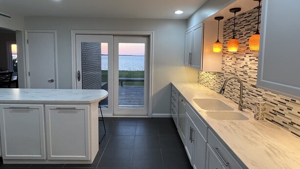 Cocina moderna con una isla grande, suelo de baldosas oscuras y vista al agua a través de una puerta de cristal.