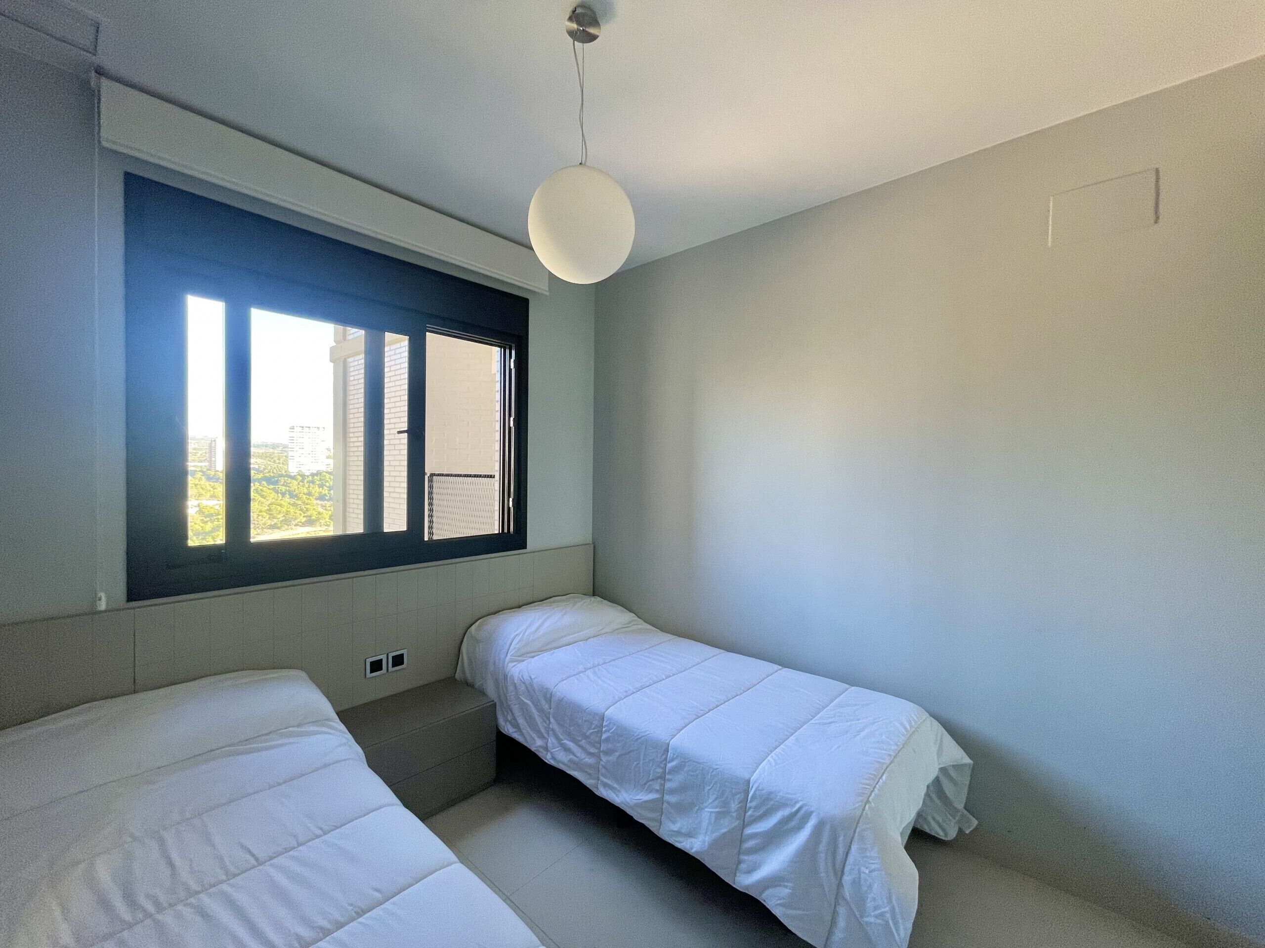 A small, modern room with two single beds, a window with blinds, and a hanging light fixture.
