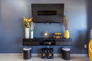 A modern living room with a flat-screen TV mounted on the wall, a black entertainment unit with two speakers, and two round stools. - Warm & nicely decorated unit @ unbeatable location (Oranjestad)