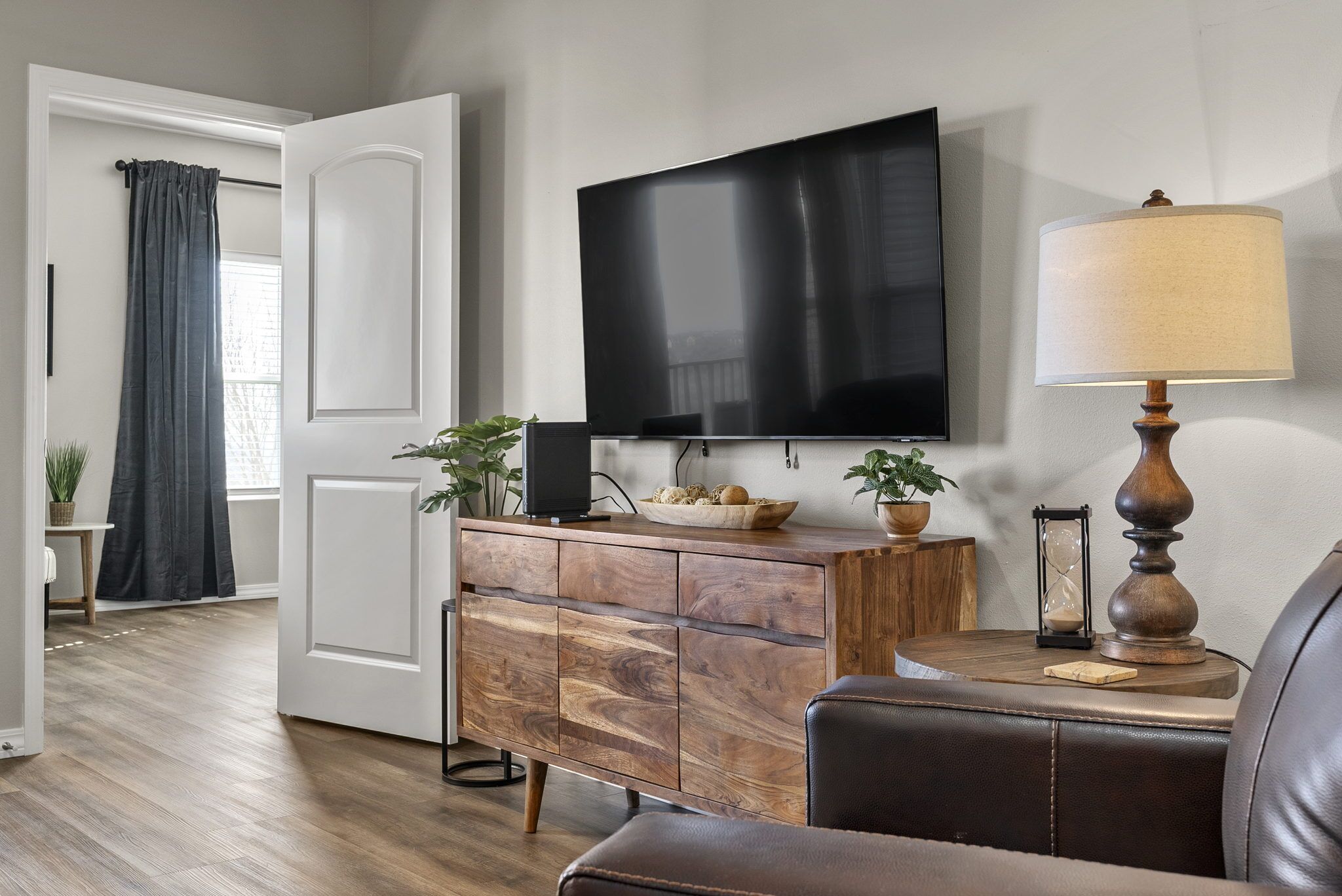 Een woonkamer met een flatscreen televisie aan de muur, een houten kast met lades, een leren bank en een bijzettafeltje met een lamp en decoratieve objecten.