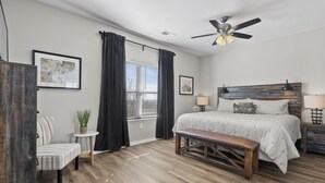 A bedroom with a wooden floor, a bed with a headboard, a bench, a chair, a small table with a plant, and a window with curtains. - Matt's Adventure Condo (Branson)