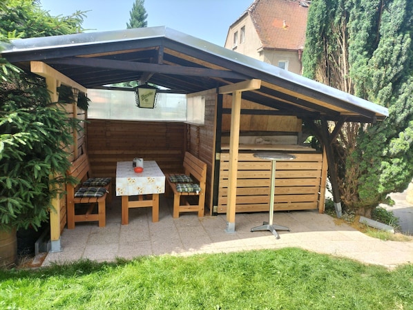 A wooden outdoor seating area with a table and benches, a grill, and a covered roof structure.
