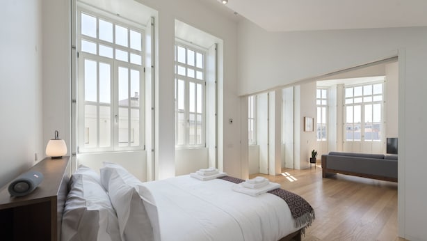 A modern bedroom with a large bed, bedside table, and a sofa in a bright room with large windows.