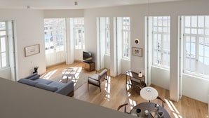 A spacious living room with a sofa, a TV, a small table, and a chair. Large windows allow natural light to fill the room.