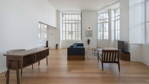 Un salón amplio con suelo de madera, un sofá, una mesa de centro de madera, un televisor y ventanas grandes.