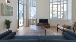 Amplia sala de estar con un sofá grande, una mesa de centro y una televisión. Hay una planta, un ventanal y vistas de edificios.