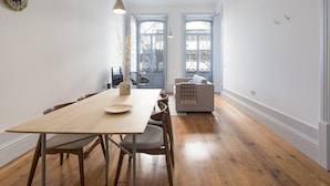 Uma sala de jantar com uma mesa e cadeiras de madeira, duas janelas com molduras azuis e uma parede branca.