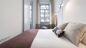 Quarto de hotel moderno com uma cama grande, janela com cortinas, abajur e uma cadeira pequena.