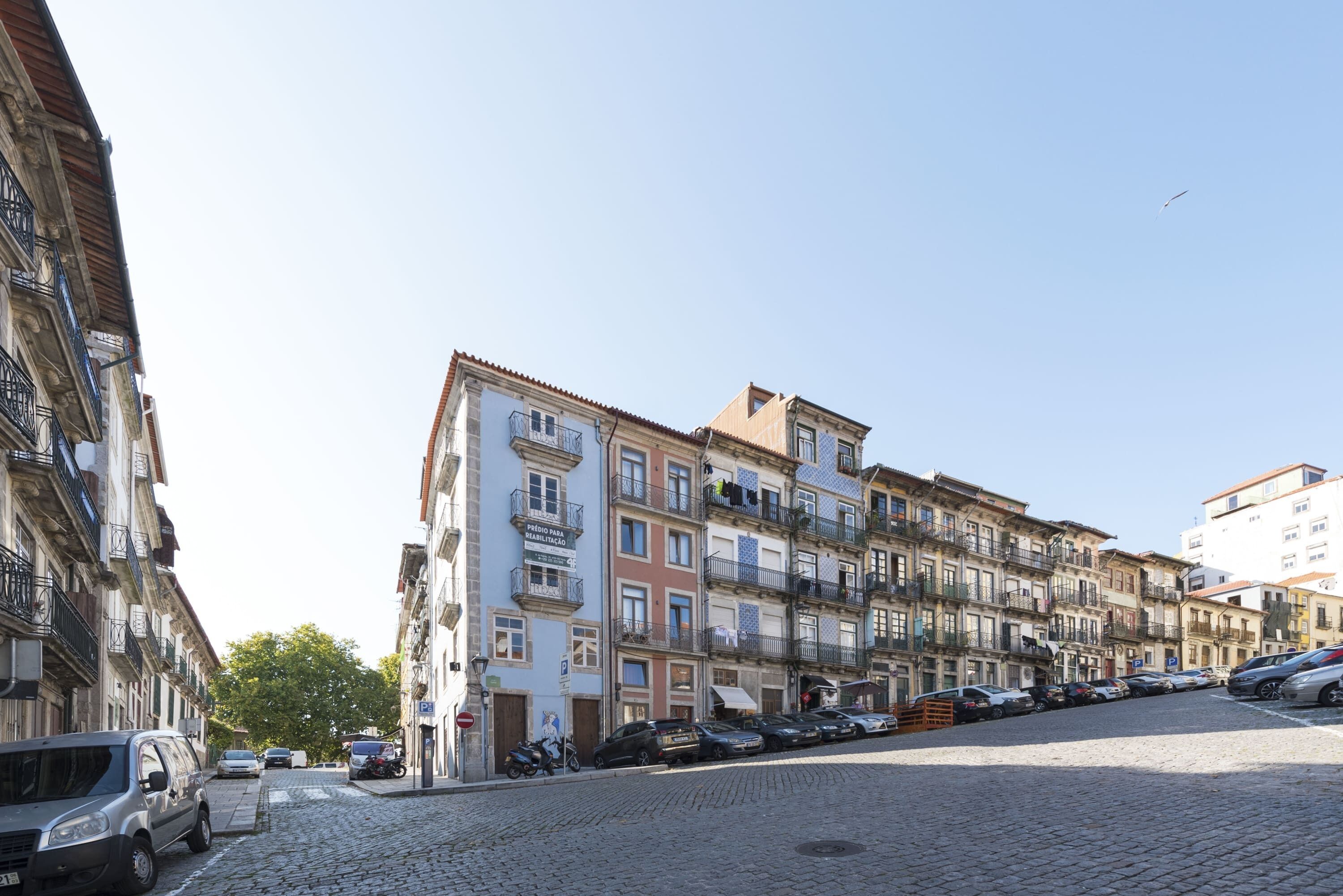 Une rue pavée bordée d’immeubles à plusieurs étages, de voitures garées et sous un ciel dégagé.