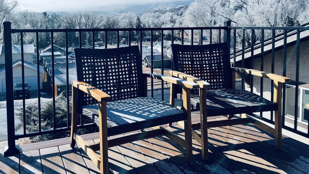 A balcony with two wooden chairs and a metal railing overlooking a snowy landscape with houses and mountains. - The Apartment Suite! Private Hot Tub w/ Mountain Views (Colorado Springs)