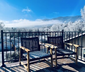 Een balkon met twee houten stoelen en een metalen hekwerk met uitzicht op een besneeuwd landschap met huizen en bergen.