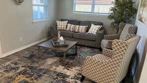 Ein Wohnzimmer mit grauem Sofa, gemusterten Sesseln, einem Couchtisch und einer großen Pflanze.