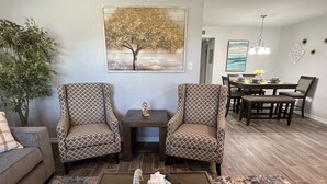 Ein Wohnzimmer mit zwei gemusterten Sesseln, einem Couchtisch mit Fernbedienung und Taschentüchern, einer Essecke mit Tisch und Stühlen sowie einem Baumgemälde an der Wand.