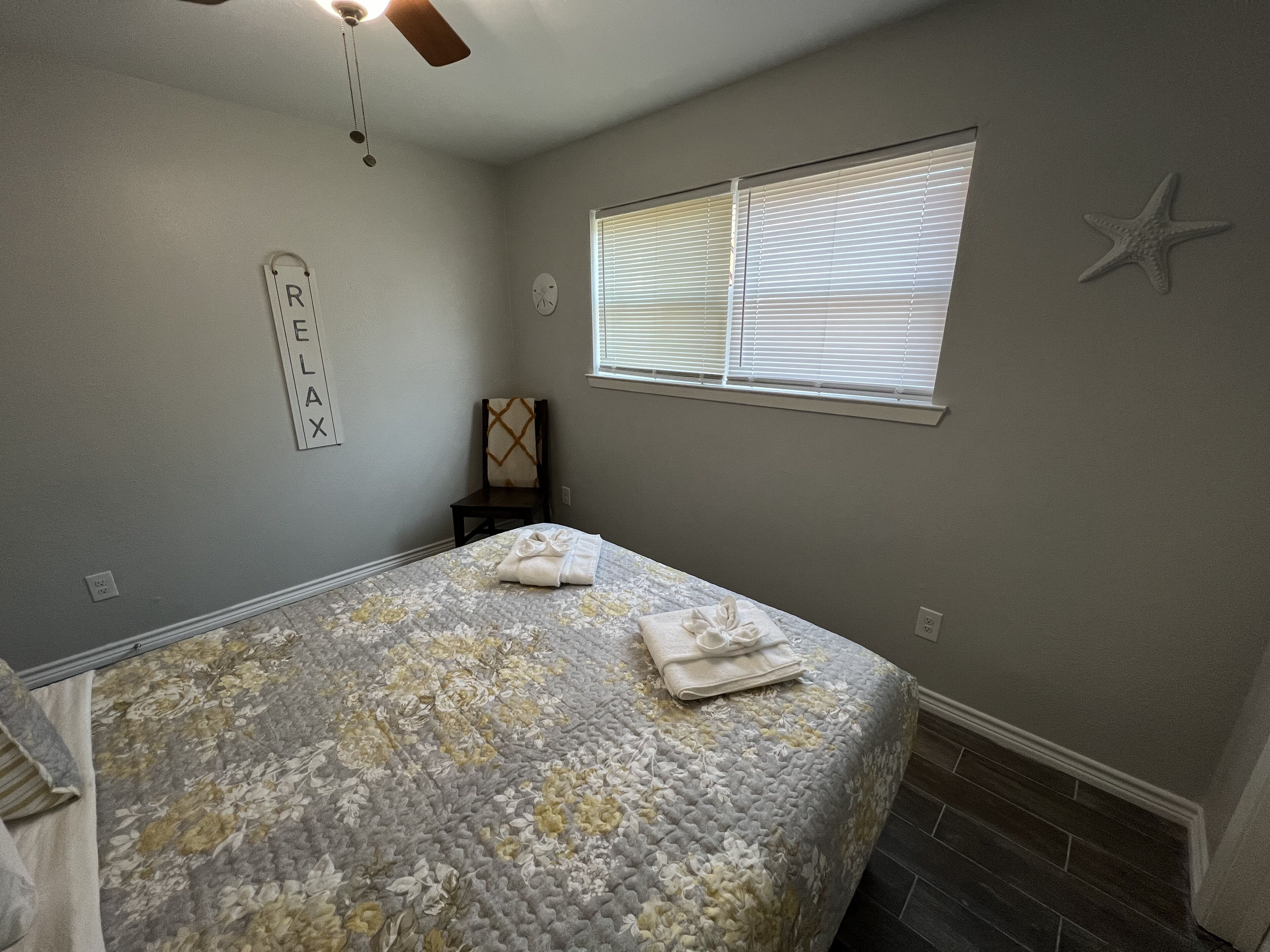 A bedroom with a bed, a ceiling fan, a window with blinds, a starfish decoration, and a wall-mounted clock.