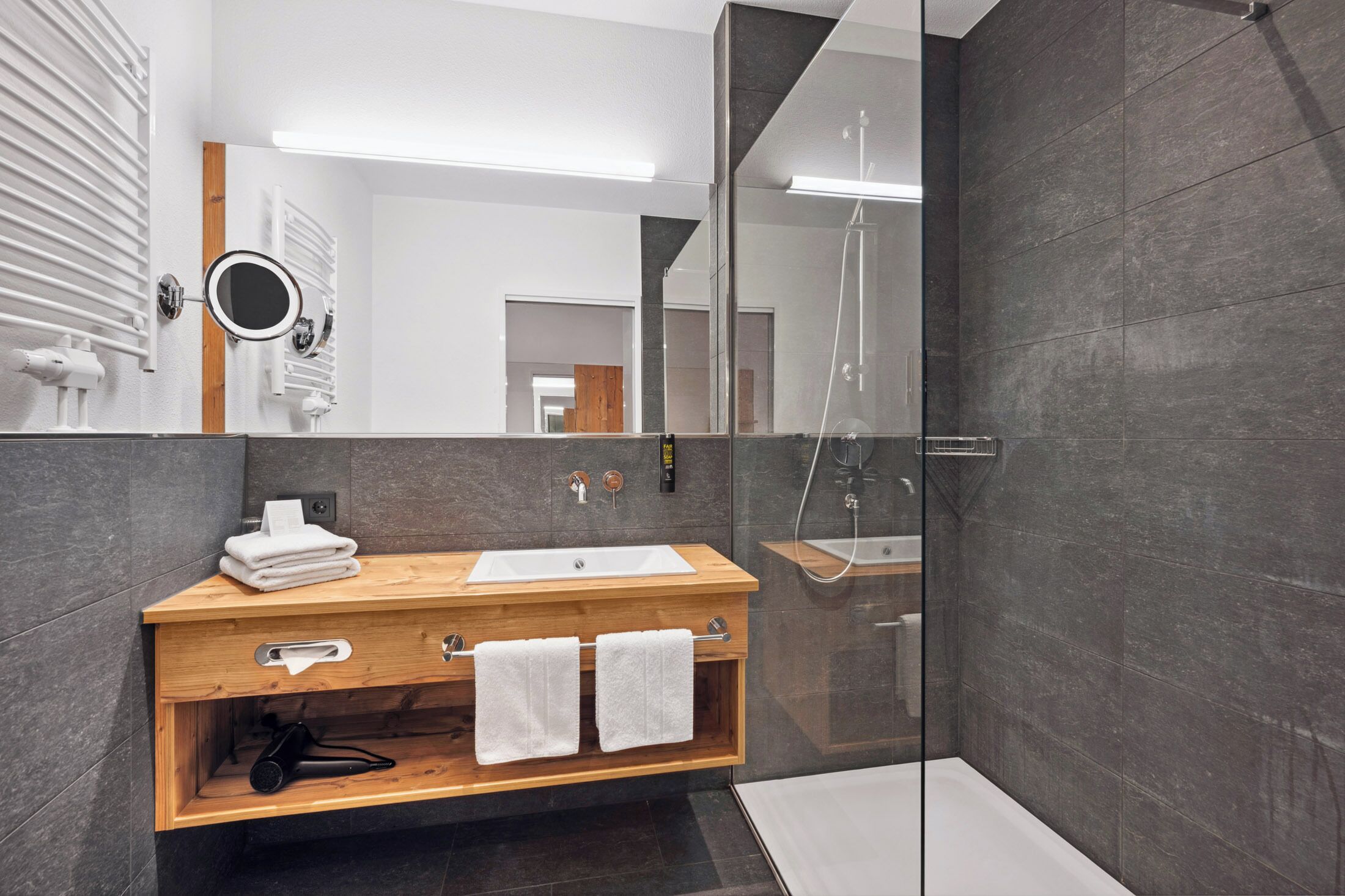 Ein modernes Badezimmer mit einem Holzmöbel, einem großen Spiegel, einer Duschkabine aus Glas und dunkel gefliesten Wänden.