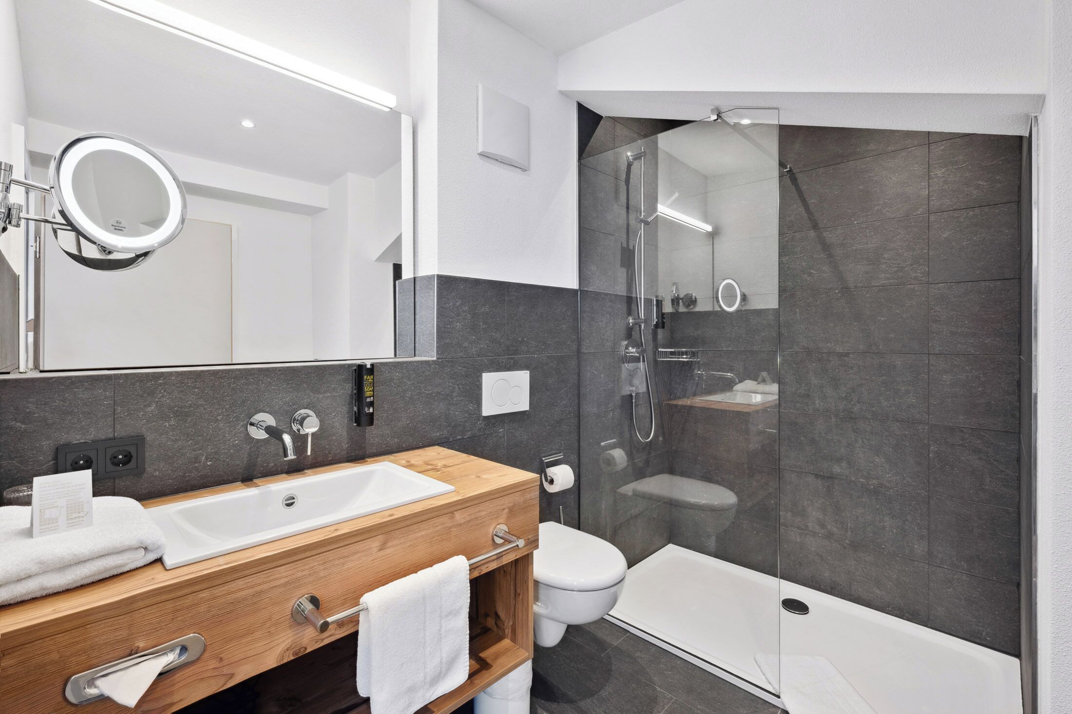 A modern bathroom with a wooden vanity, a large mirror, a white sink, a toilet, and a glass-enclosed shower area.