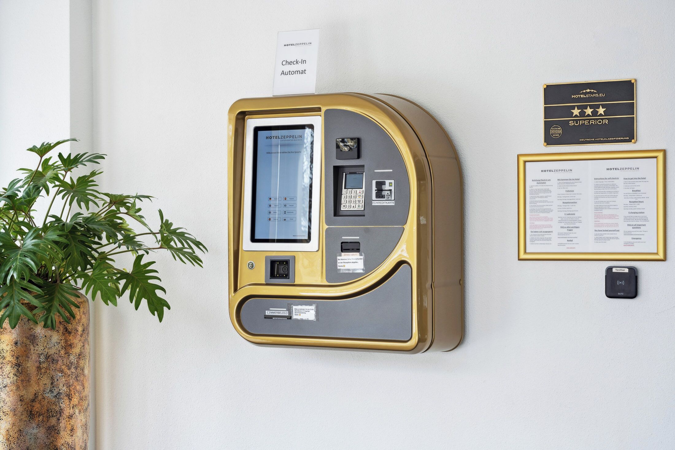 Ein Check-in-Terminal mit digitalem Bildschirm, Tastatur und kleinem Display. Eine Plakette kennzeichnet eine 'Superior'-Bewertung und eine gerahmte Speisekarte ist an der Wand angebracht.