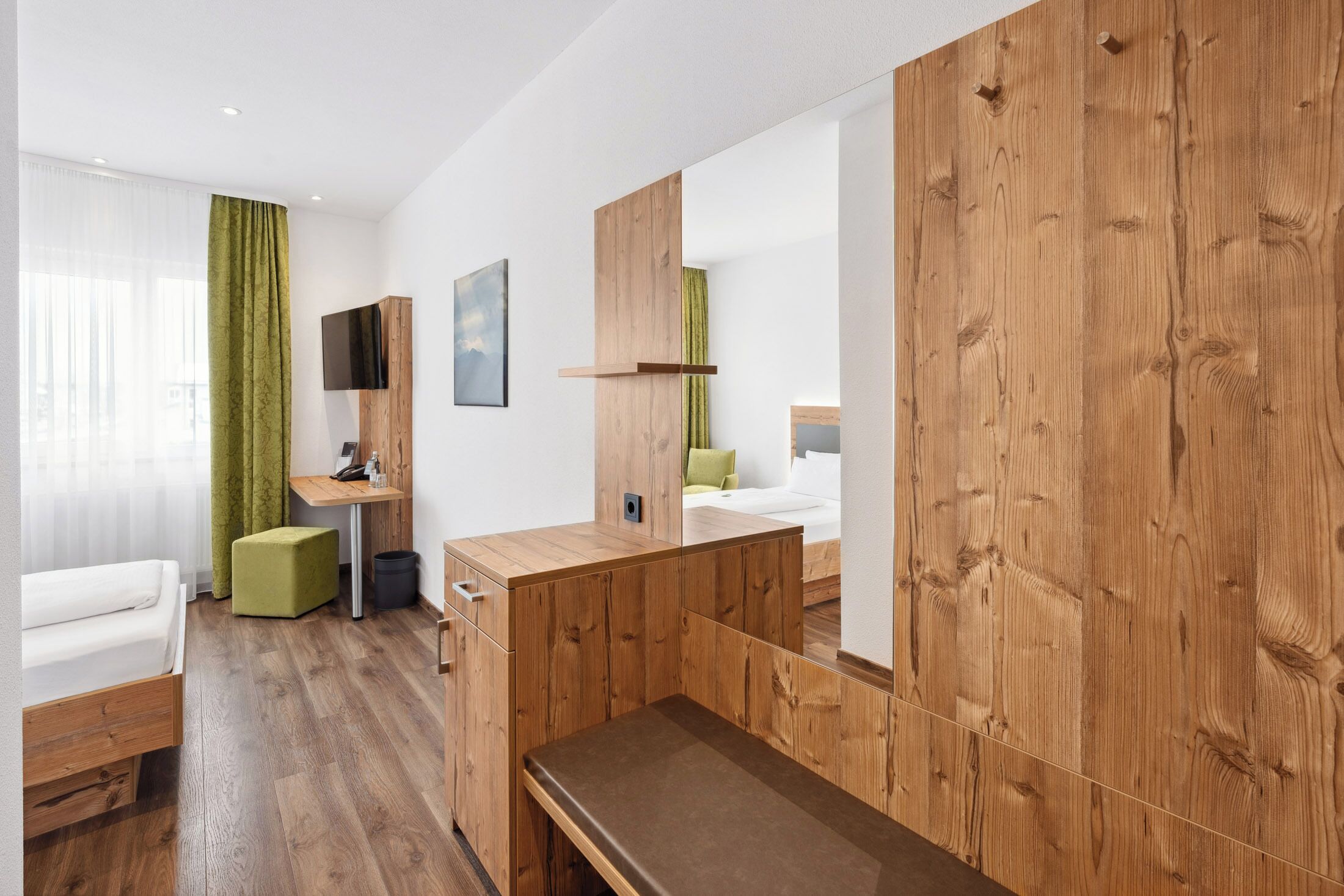Ein modernes Hotelzimmer mit einem Holztisch, einem Bett, einem grünen Hocker und einem großen Spiegel.