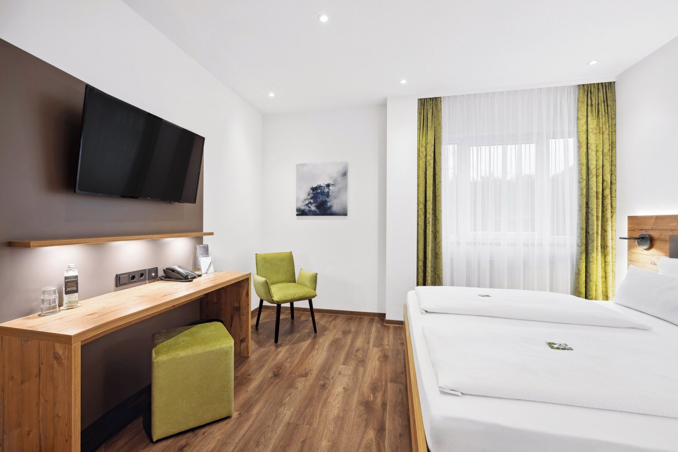 A hotel room with a bed, a desk with a chair, a television mounted on the wall, and a window with sheer curtains.