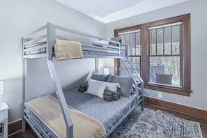 A bunk bed with a ladder, a window with blinds, a rug, and a small bedside table.