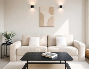A living room with a beige sofa, two wall-mounted lights, a framed abstract painting, a black coffee table with a book, and a vase with flowers on a side table. - Champs-Élysées studio with balcony & AC (Paris)