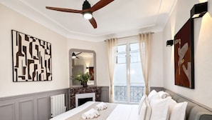 A hotel room with a bed, a ceiling fan, a fireplace, a window with curtains, and artwork on the walls.