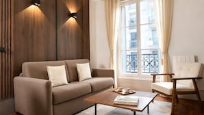 A modern living room with a sofa, a coffee table, and a chair. There is a window with curtains and a view of buildings.