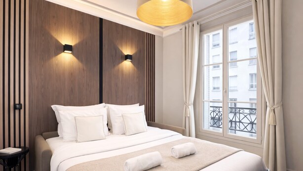 A hotel room with a bed, two wall-mounted lights, a window with curtains, and a view of buildings outside. - Charming flat with lift - Eiffel Tower (Paris)