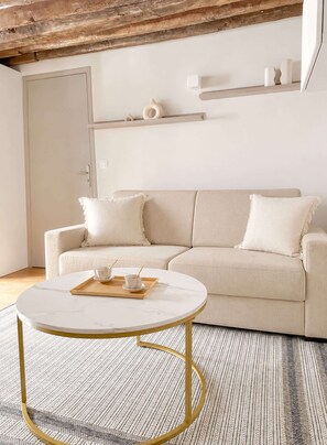A minimalist living room with a beige sofa, a round coffee table, and a wooden floor. - Cozy apartment near Palais Garnier (Paris)