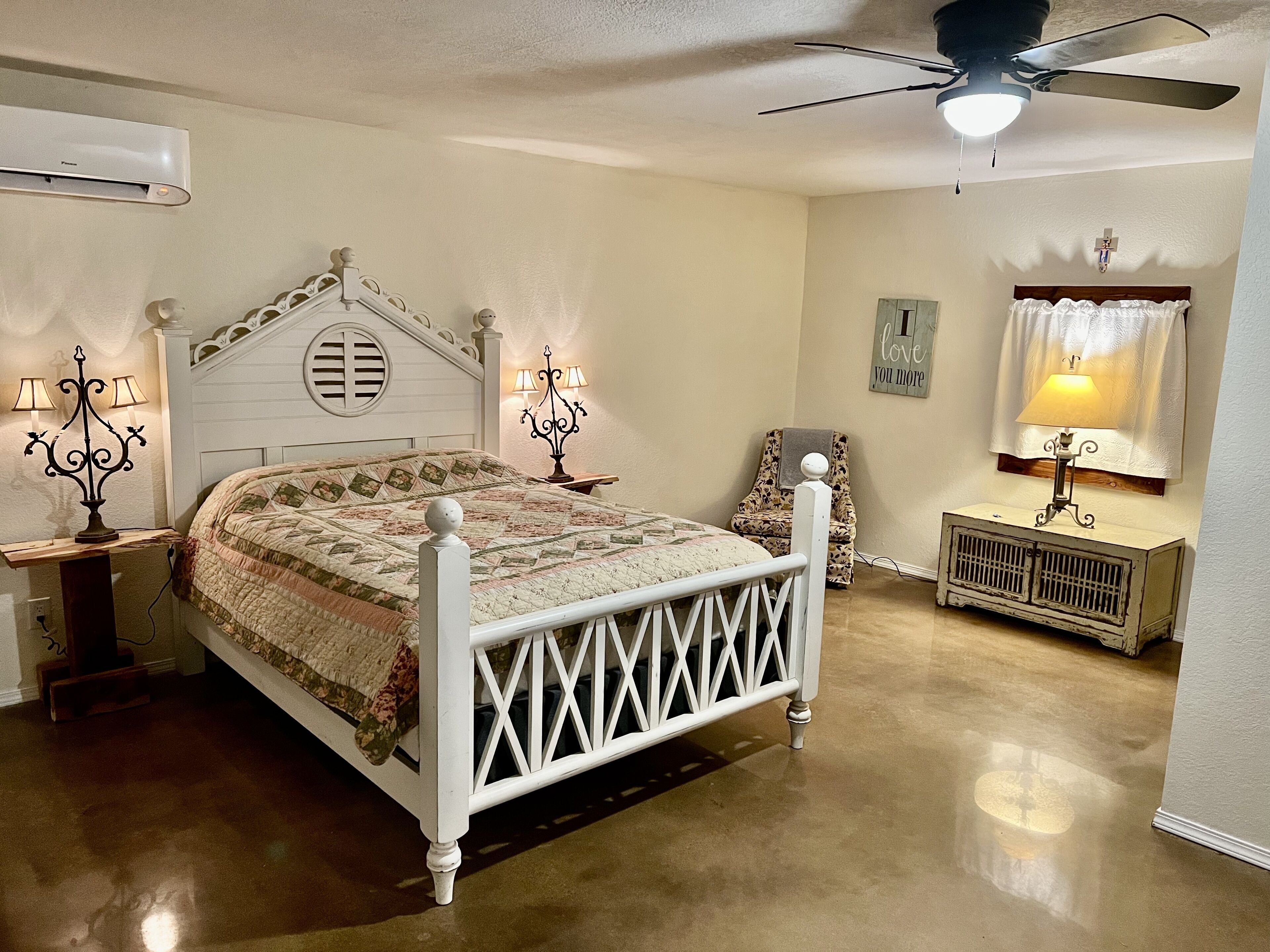 A bedroom with a white bed, a ceiling fan, a wall-mounted air conditioner, and a bedside table with a lamp.