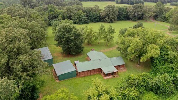 A rustic wooden cabin with a green roof, surrounded by dense forest and open fields.