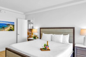 A hotel room with a bed, a nightstand, a lamp, a painting of a beach, and a bottle of champagne on the bed.
