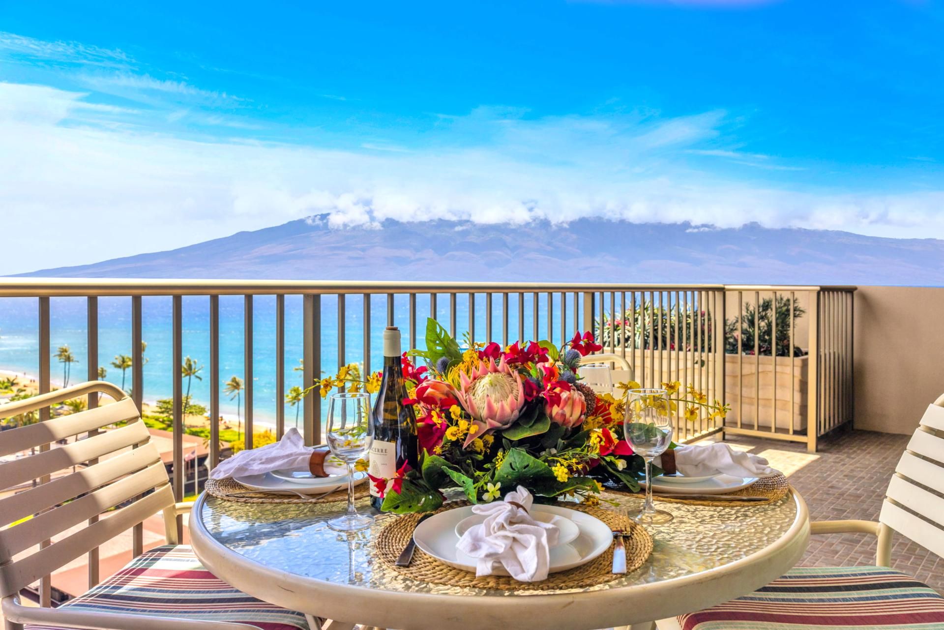 A table set for two with a bottle of wine, a glass, and a floral centerpiece on a balcony overlooking a beach and a mountain.