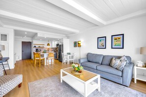 Ein modernes Wohnzimmer mit einem grauen Sofa, einem Holztisch und einem Essbereich, im Hintergrund ist eine Küche zu sehen.