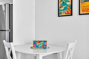 A modern kitchen with a stainless steel refrigerator, a white dining table with two white chairs, and a framed Hawaiian-themed artwork on the wall.