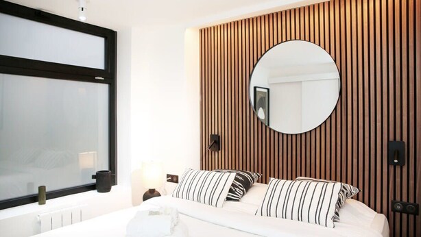 Una habitación de hotel moderna con una cama grande, un espejo redondo, paneles de madera y una ventana con persianas.