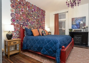 A bedroom with a large, colorful crochet wall art, a red bed frame, and a blue quilted bedspread.