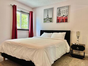 Un dormitorio con cama, una ventana con cortinas rojas, una mesita de noche con lámpara y dos cuadros colgados en la pared.
