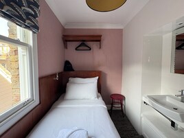 Ein kleines, schmales Hotelzimmer mit einem Einzelbett, einem roten Hocker, einem Holz-Kopfteil, einer gestreiften Wand und einem Fenster mit Blick auf Gebäude.