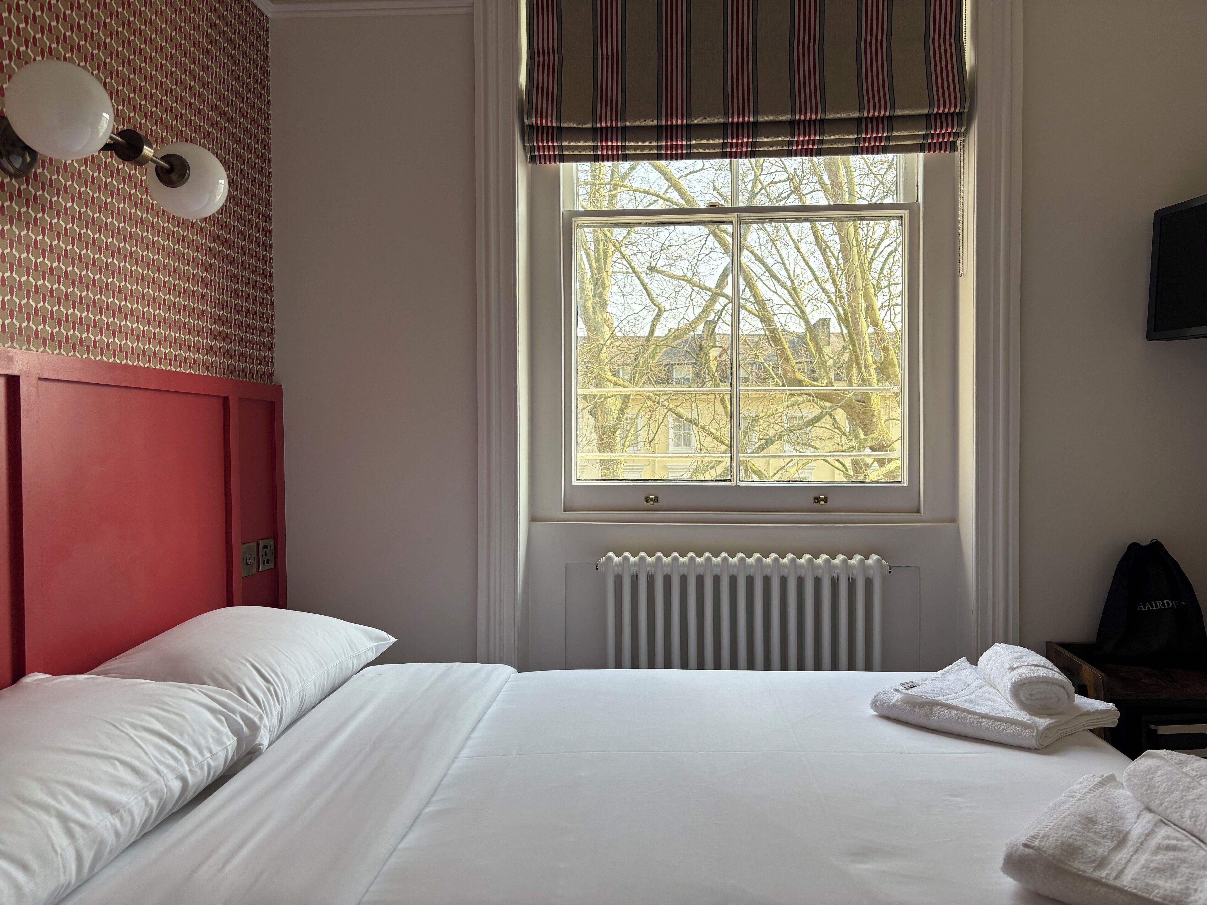 A hotel room with a bed, a radiator, a window with a view of trees, and a ceiling fan.