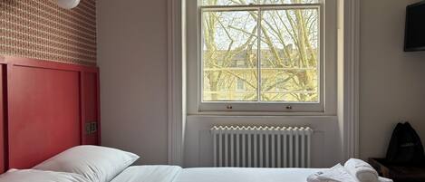 A hotel room with a bed, a radiator, a window with a view of trees, and a ceiling fan.