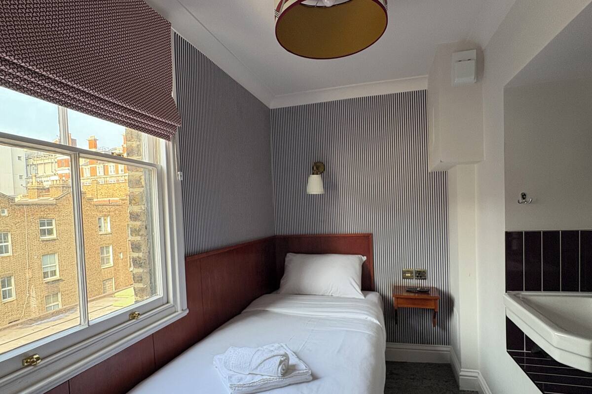 A hotel room with a single bed, a small bedside table, a nightstand, a window with a view of buildings, and a round ceiling light.