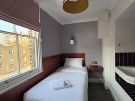 Ein Hotelzimmer mit einem Einzelbett, einem kleinen Nachttisch, einer Lampe und einem Fenster mit Blick auf Gebäude. 