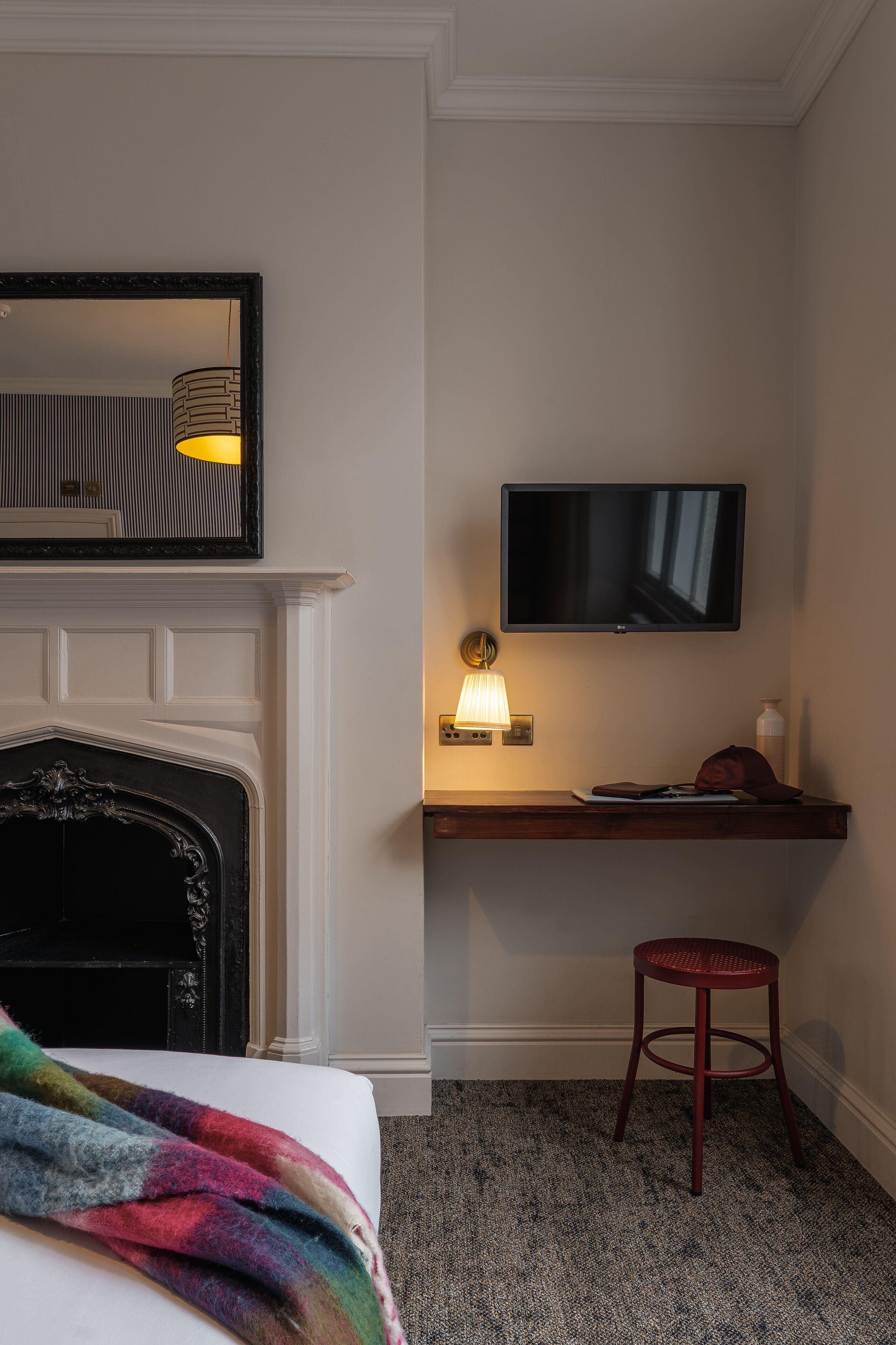 A hotel room with a fireplace, a television mounted on the wall, a desk with a lamp, and a red stool.