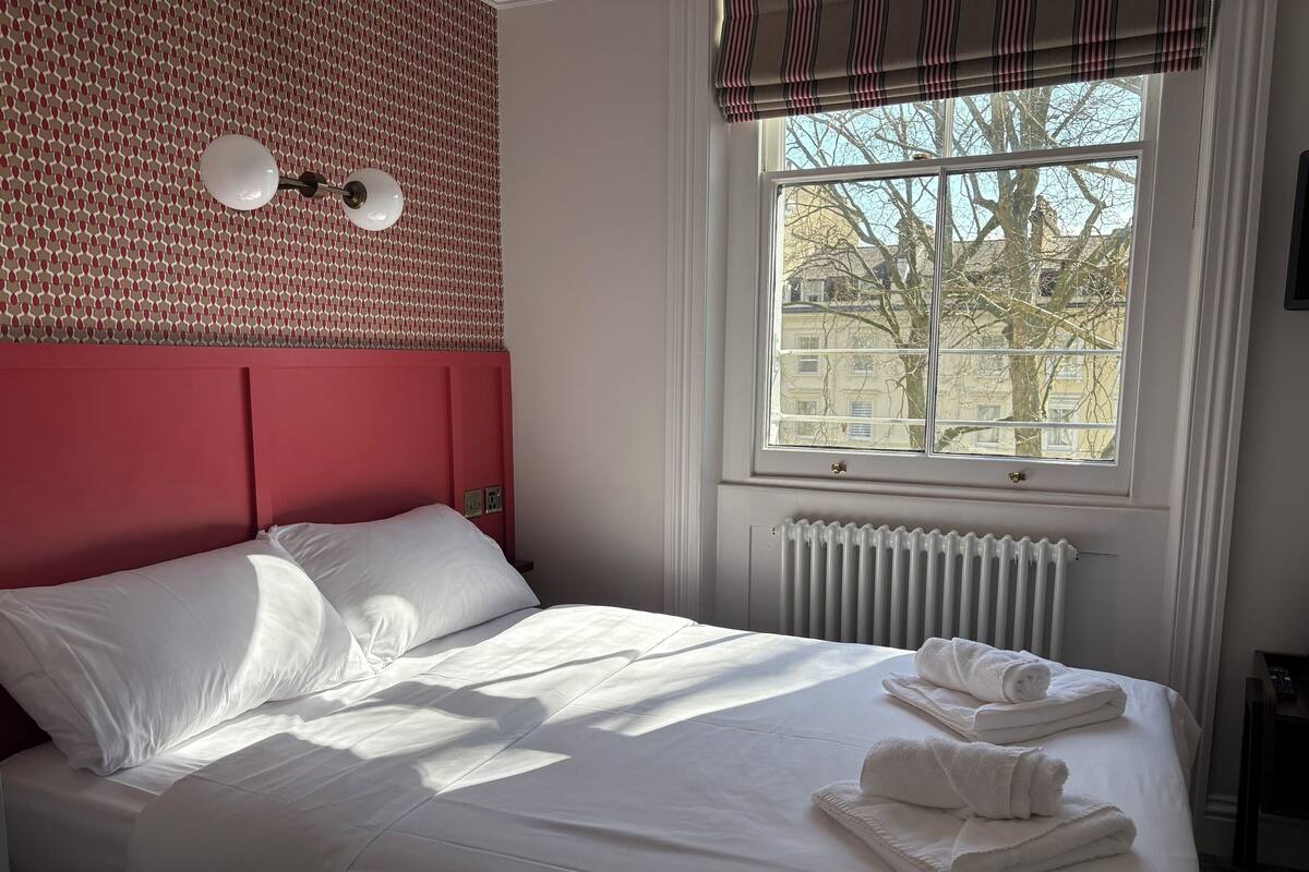 A neatly made bed with white linens and towels, a red headboard, a window with a view of buildings, and a radiator below the window.