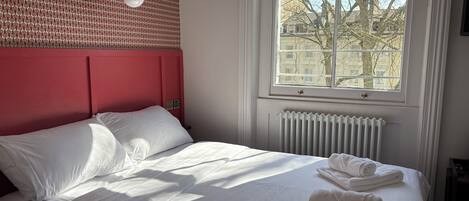 Een opgemaakt bed met witte lakens en handdoeken, een rood hoofdeinde, een raam met uitzicht op gebouwen en een radiator onder het raam.