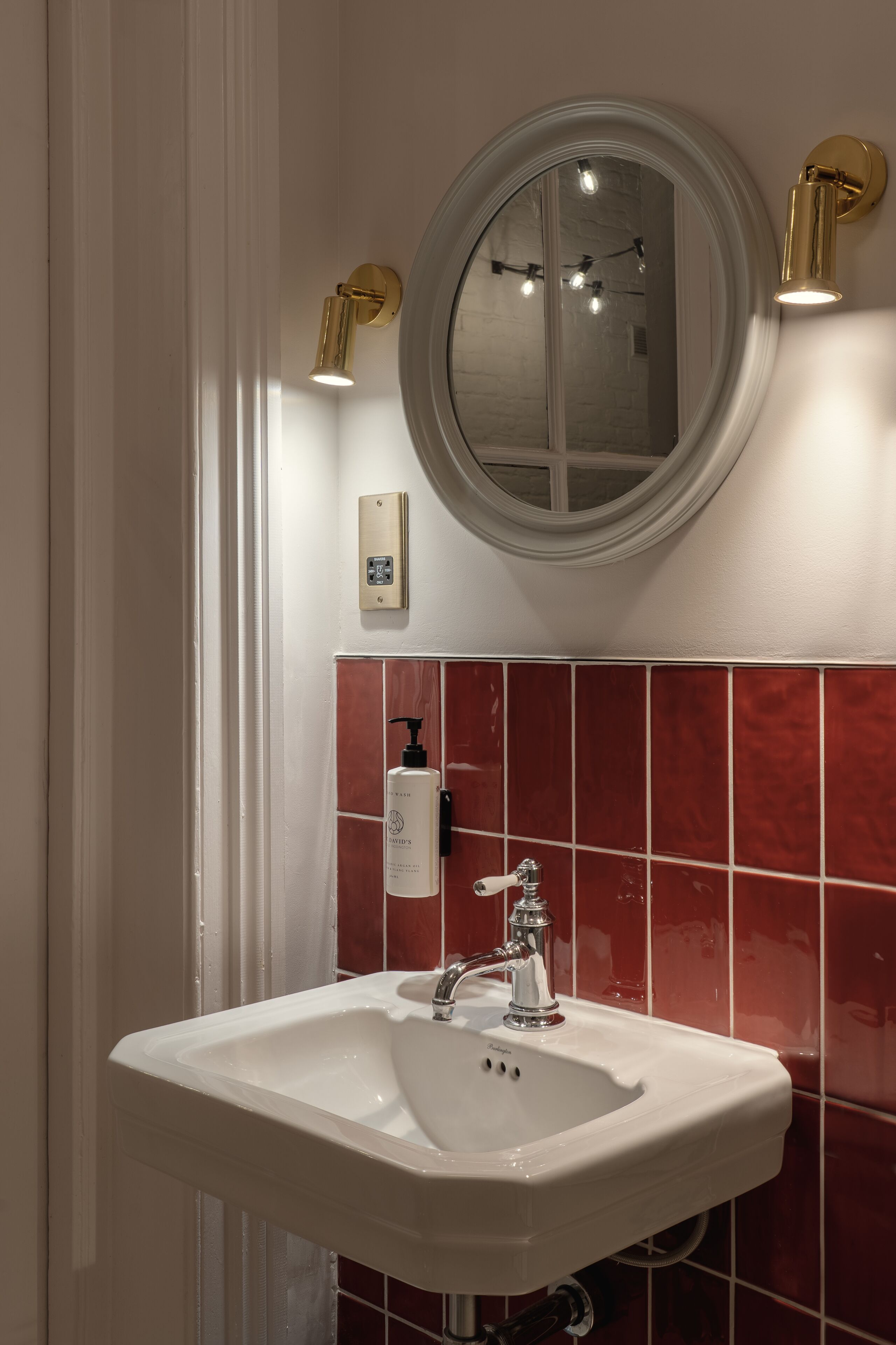 A bathroom with a white sink, red-tiled walls, and a round mirror with two wall-mounted lights.