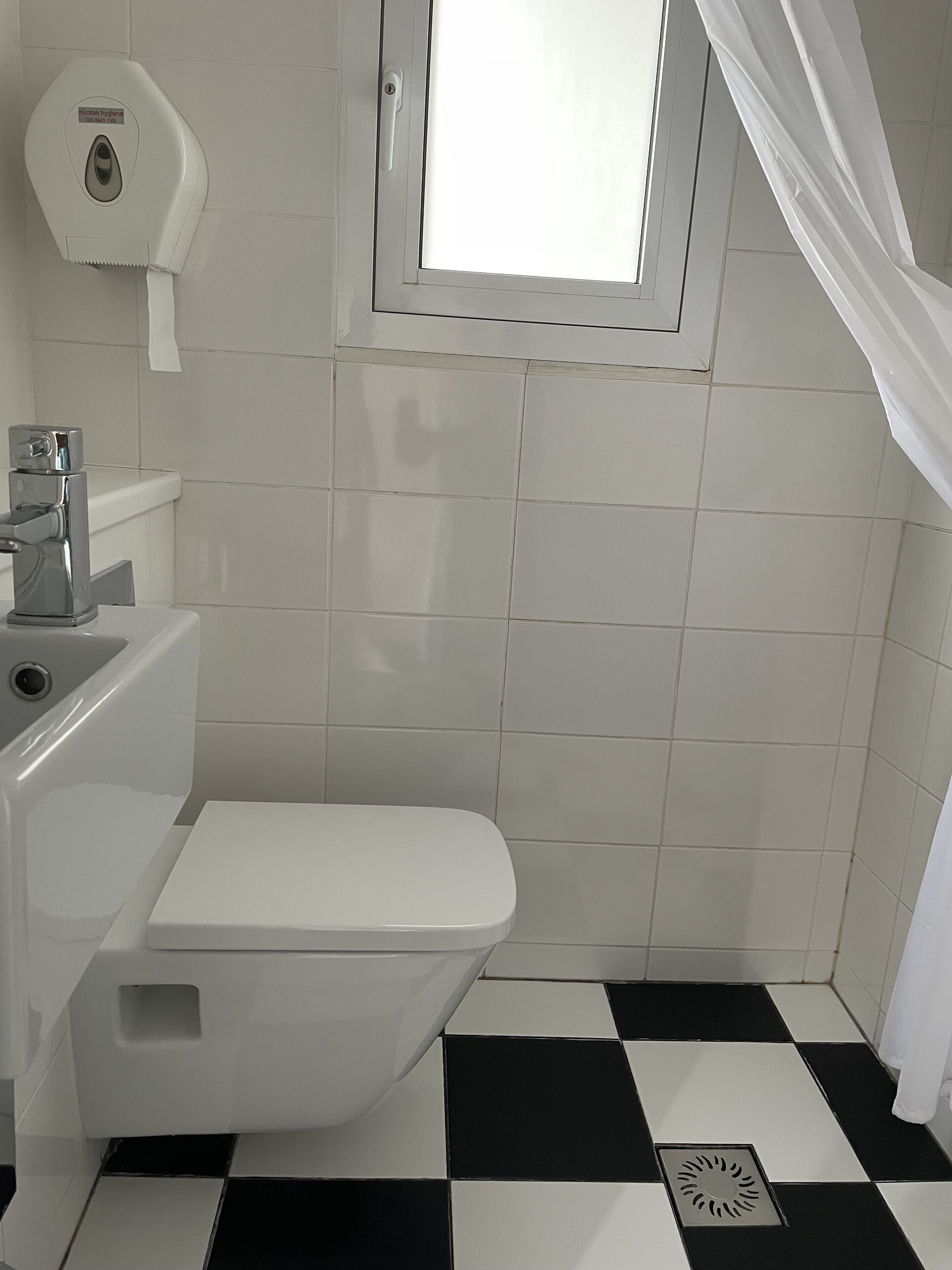 A bathroom with a toilet, a hand dryer, and a window with a curtain.