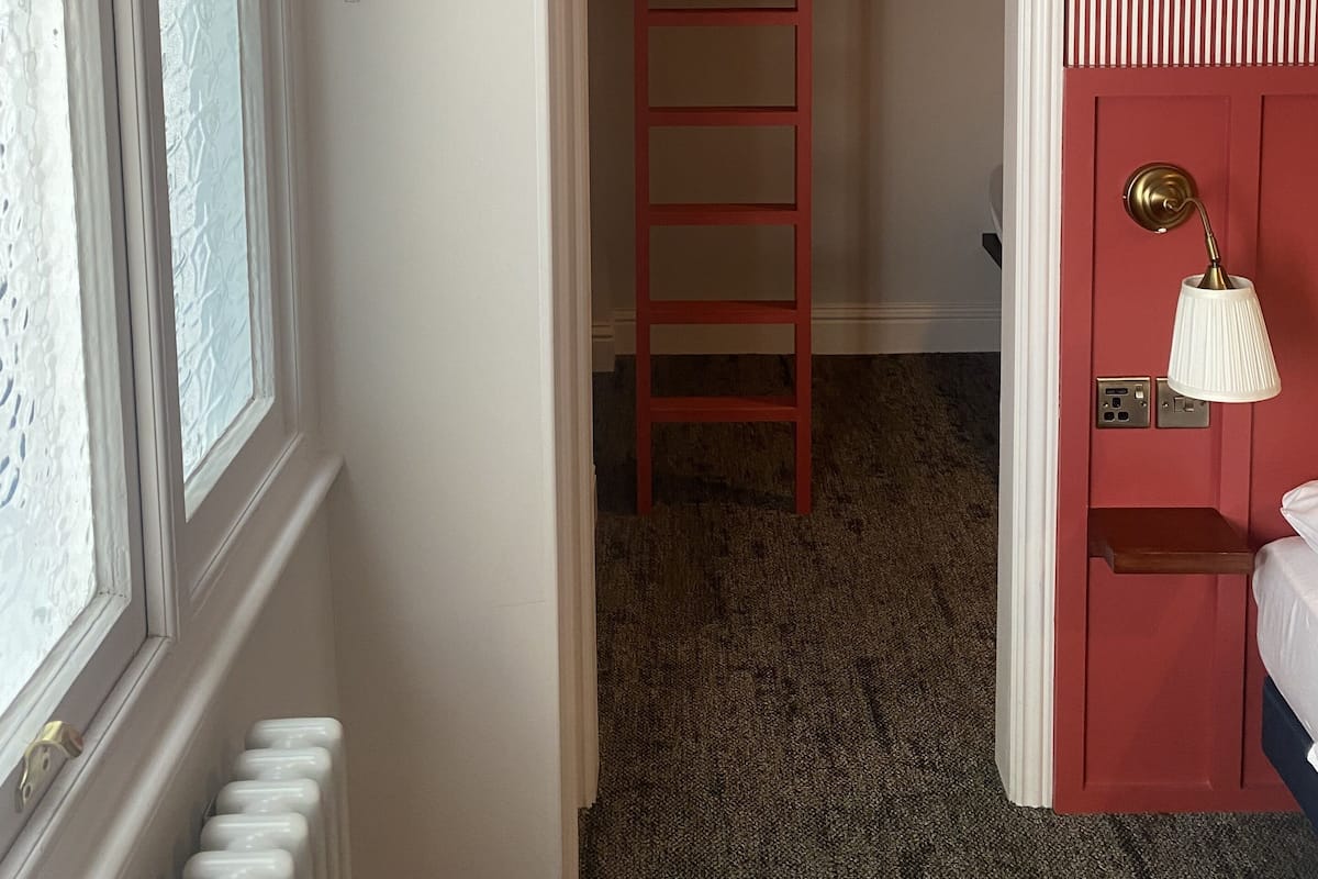 A room with a bunk bed, a window with a patterned curtain, a radiator, and a wall-mounted lamp.